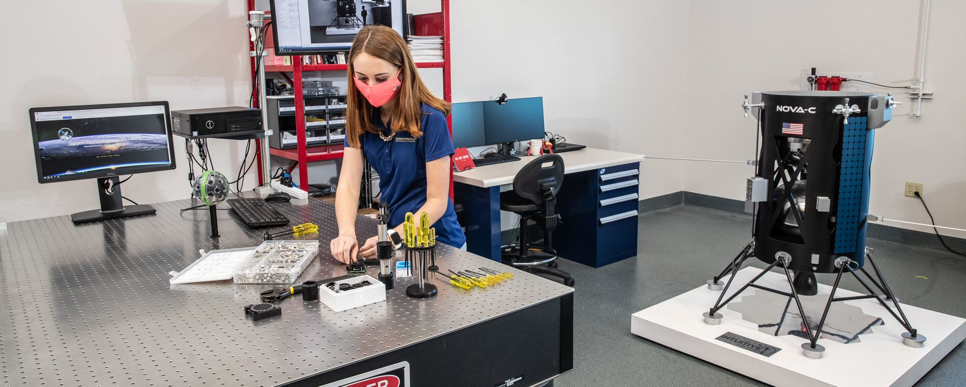 Taylor Yow, an Aerospace Engineering undergrad, examines a camera lens at the optical table in the Space Technologies Lab at Embry-Riddle Aeronautical University. The Niceville, Florida, resident works on EagleCam&rsquo;s sensing team and oversees project management. EagleCam will be capturing images of the Nova-C Lunar Lander (early model pictured in foreground) as it touches down on the Moon in fall 2021.
