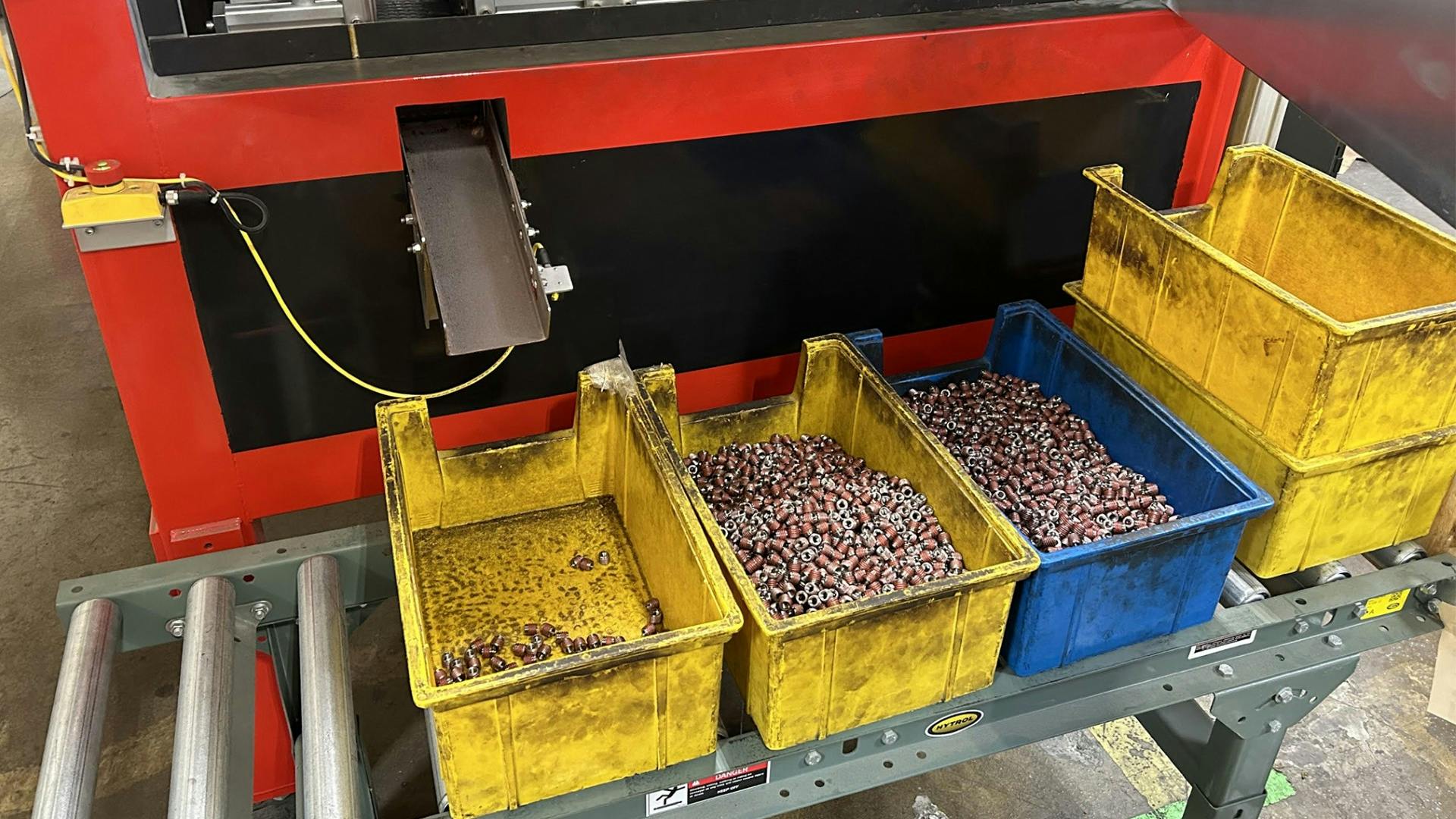 A group of yellow and blue plastic bins filled with coated metal threaded inserts on a conveyor belt