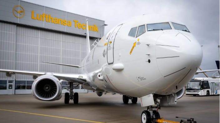 A white airplane sits in front of a grey building with yellow letters that read, 'Lufthansa Technik'