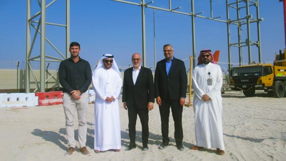 Five men in professional attire stand smiling at the camera on a construction site, with metal framework of a building behind them