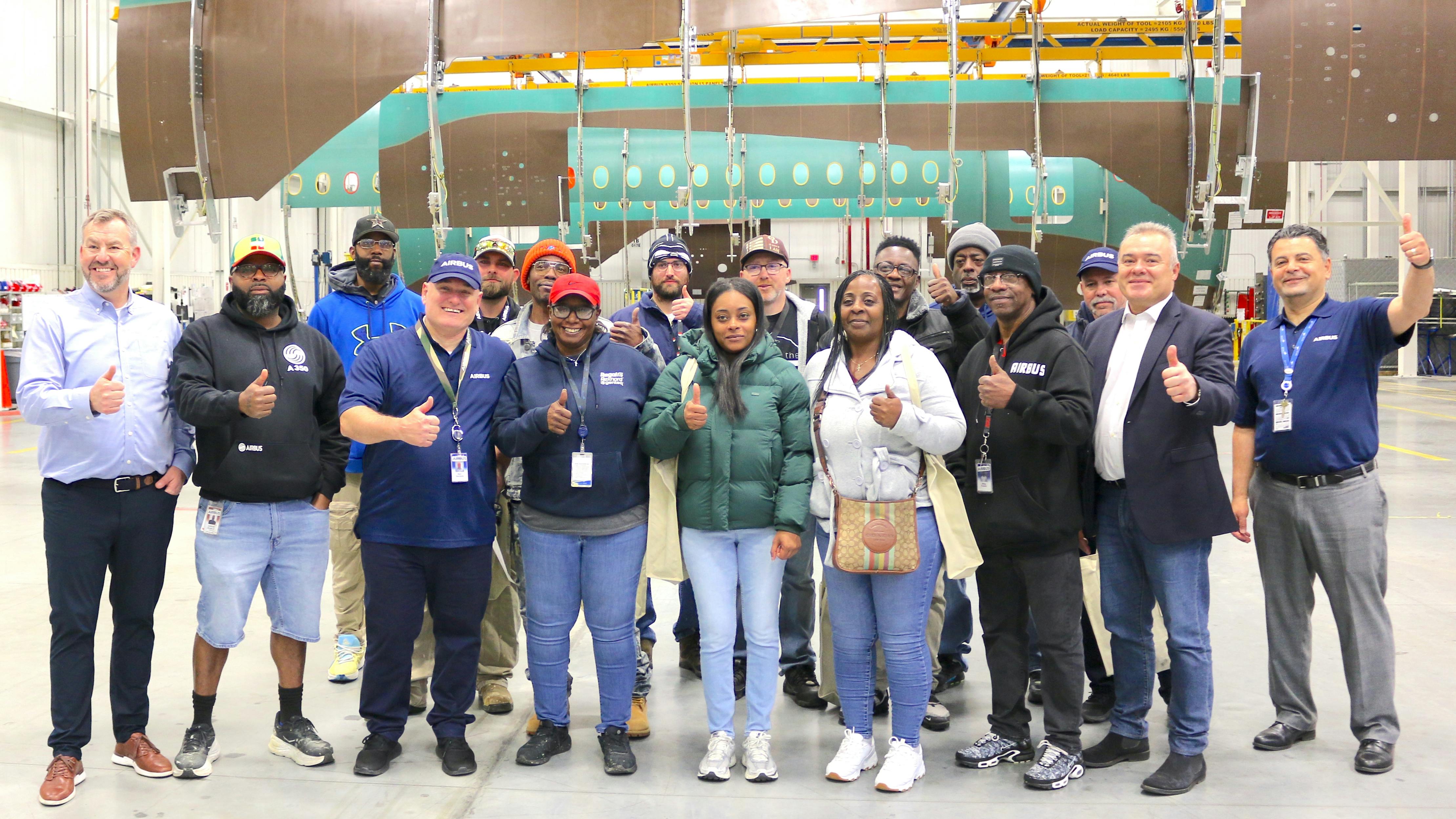 A group of employees stand in an aircraft hangar in front of a deconstructed airplane, giving thumbs-ups and smiling at the camera