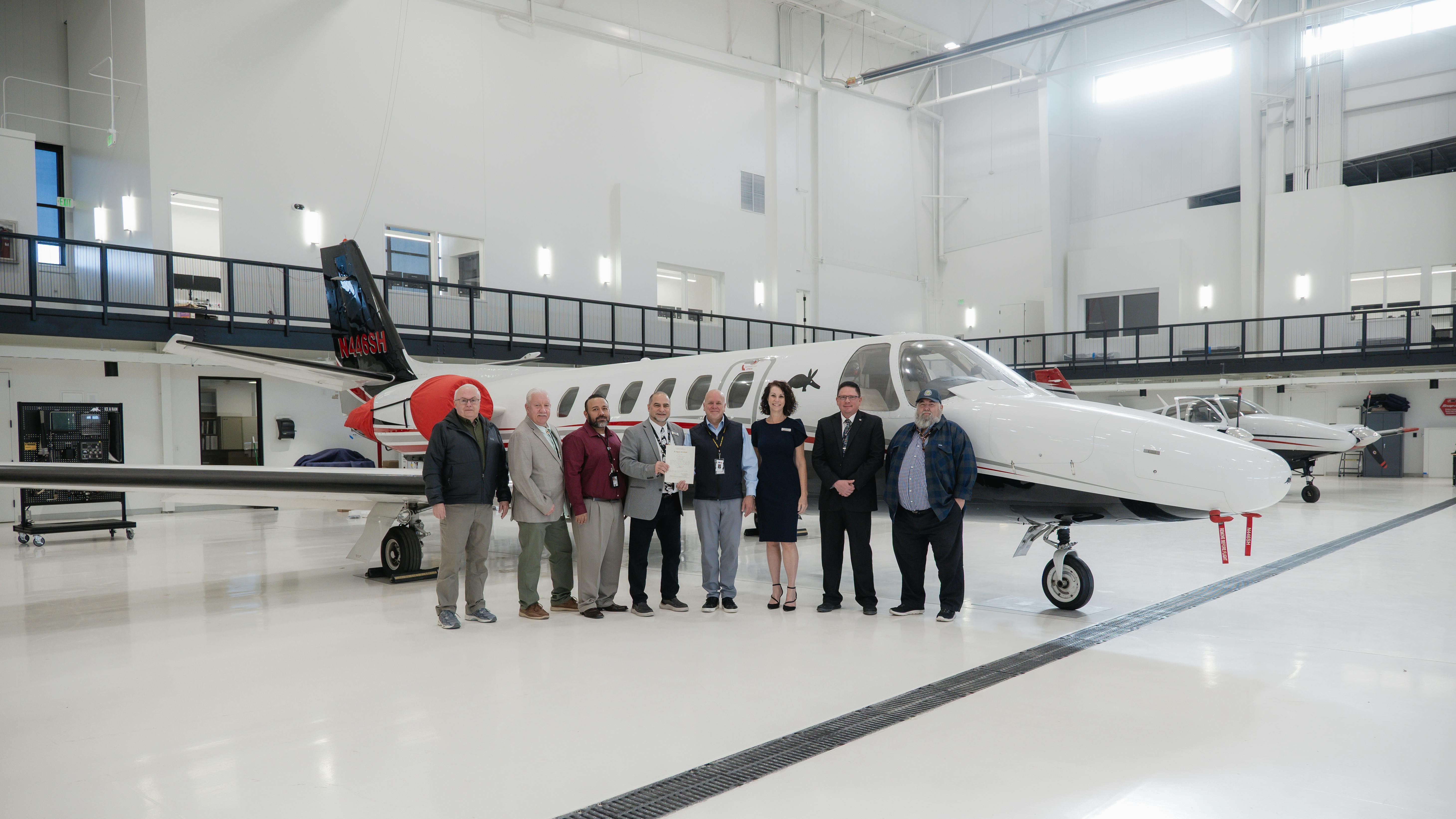 A group of professionals in business attire stand in front of an airplane in an airplane hangar, smiling at the camera