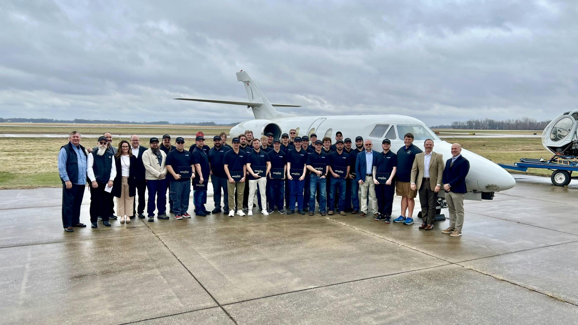 A group of professionals in business attire and AMT students in West Star Aviation Academy polos pose together in front of a white airplane