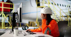 A female maintenance technician wearing orange high-visibility clothing and a white hard hat sits at a computer, lifting her hand to her face in a frustrated gesture A female maintenance technician wearing orange high-visibility clothing and a white hard hat sits at a computer, lifting her hand to her face in a frustrated gesture
