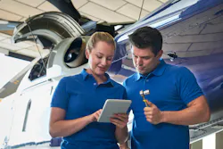 Two aircraft maintenance technicians wearing blue polos stand in front of an airplane looking at a mobile tablet, with one technician holding a socket wrench Two aircraft maintenance technicians wearing blue polos stand in front of an airplane looking at a mobile tablet, with one technician holding a socket wrench