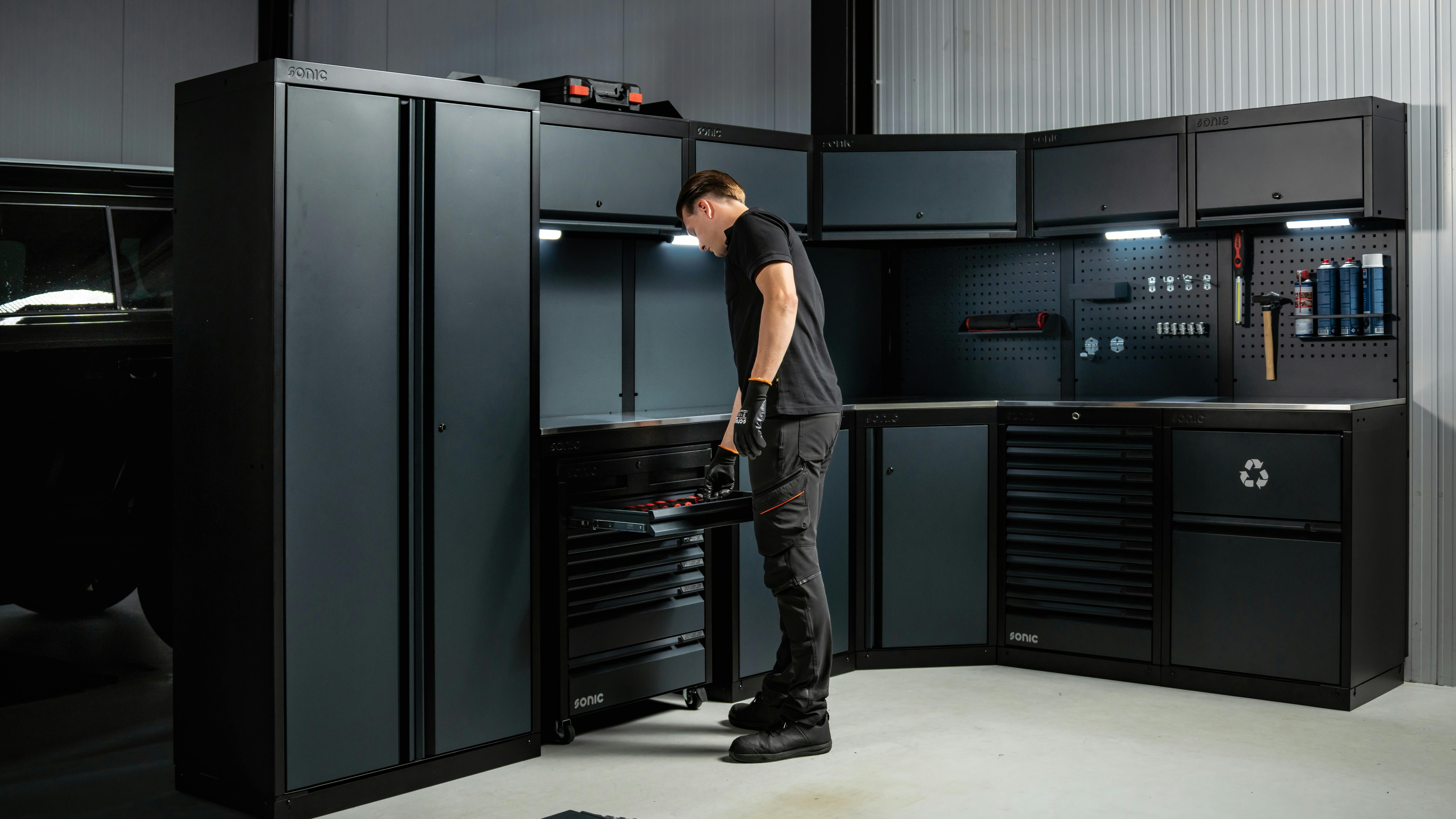 A man in black clothing standing next to a large system of black cabinets and shelves, with a tool box on the top of one of the shelves