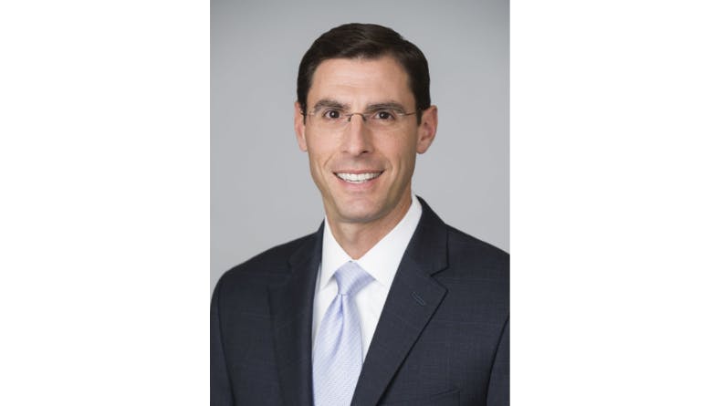 A man wearing a suit and tie and glasses smiles at the camera