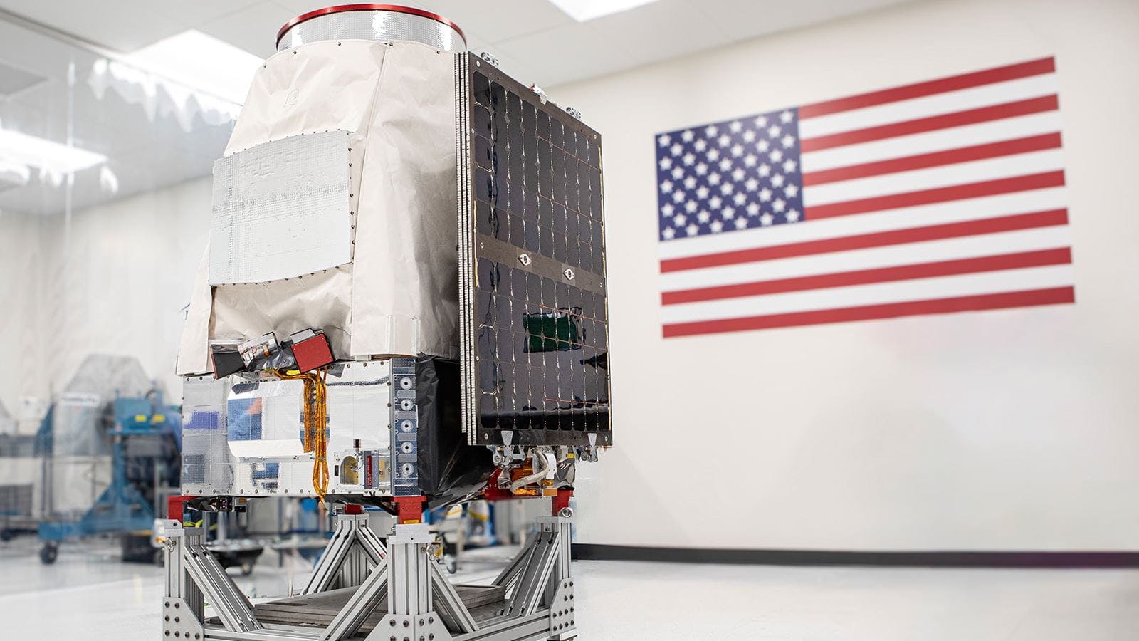 A Saturn-200 minisatellite sitting in a white room with an American flag behind it