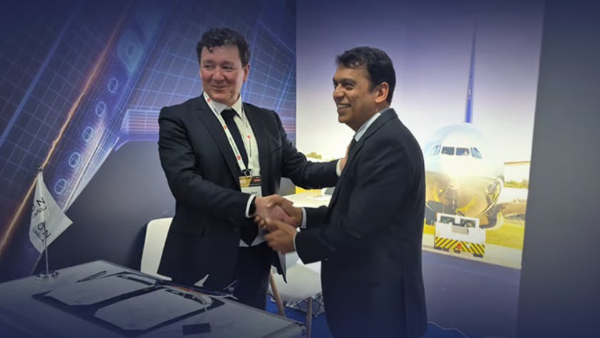 Two men wearing suits shaking hands while standing in front of a projection of an airplane, with a table in front of them holding two pieces of paper and a pen