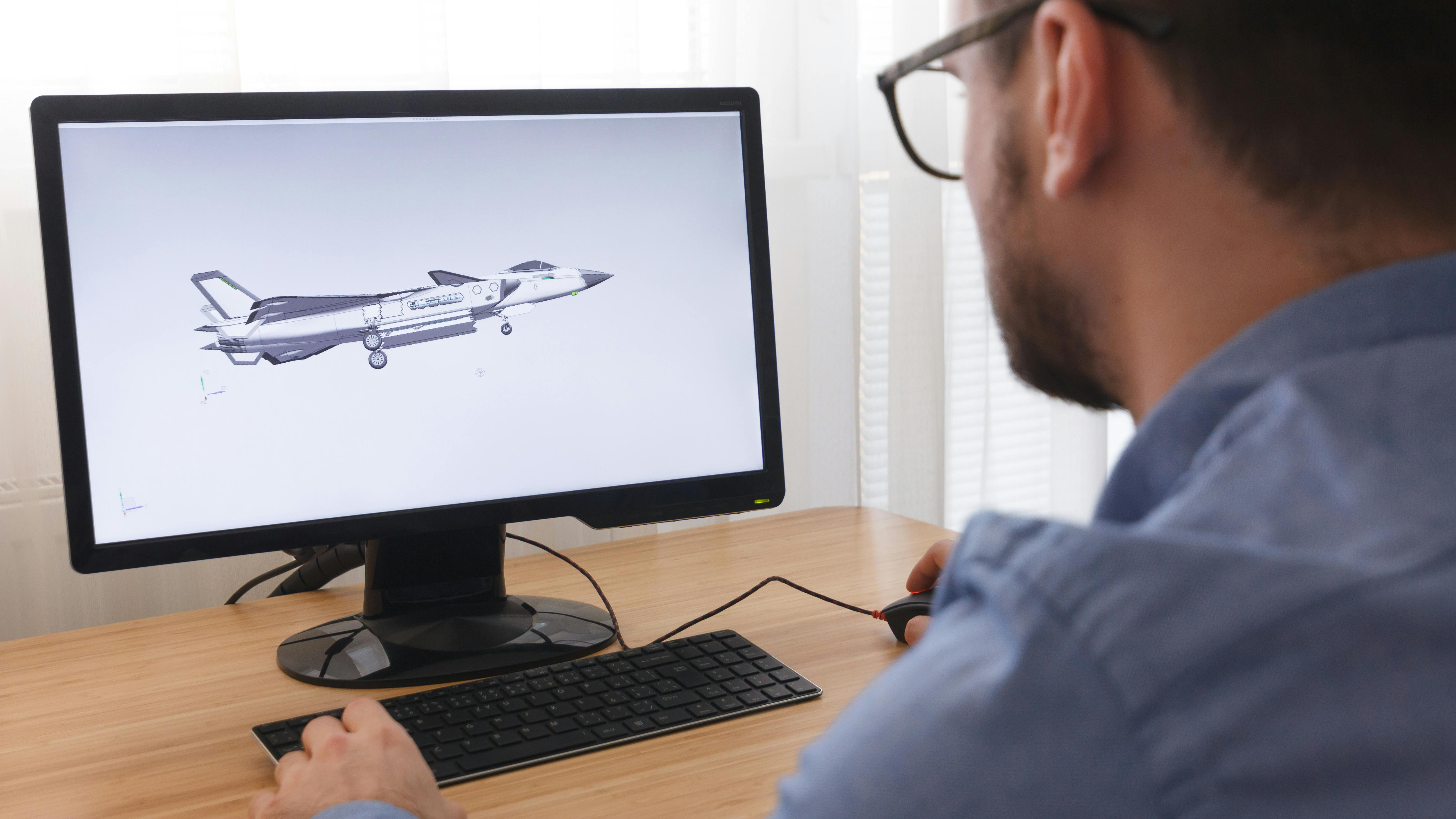 A man looking at a desktop computer that's displaying a 3D digital rendering of an airplane