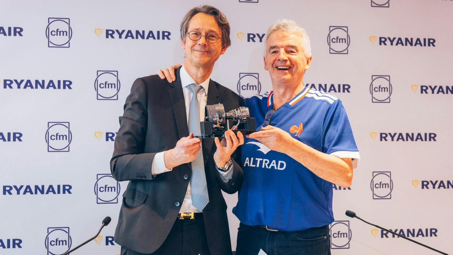 Two men standing and smiling at the camera with one holding a piece of machinery