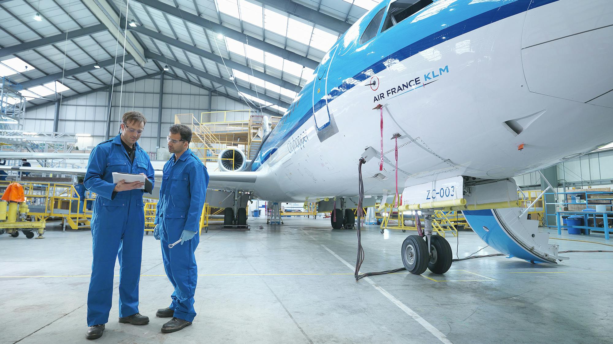 Two aircraft maintenance technicians wearing blue uniforms looking at a stack of papers while standing next to a blue and white airplane in a maintenance hangar