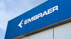 The side of a building with a beige wall and a dark blue sign with white lettering that reads: EMBRAER The side of a building with a beige wall and a dark blue sign with white lettering that reads: EMBRAER