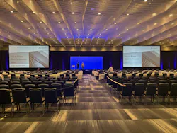 A wide shot of a room with rows of chairs in front of a stage that features a podium and projector screens A wide shot of a room with rows of chairs in front of a stage that features a podium and projector screens