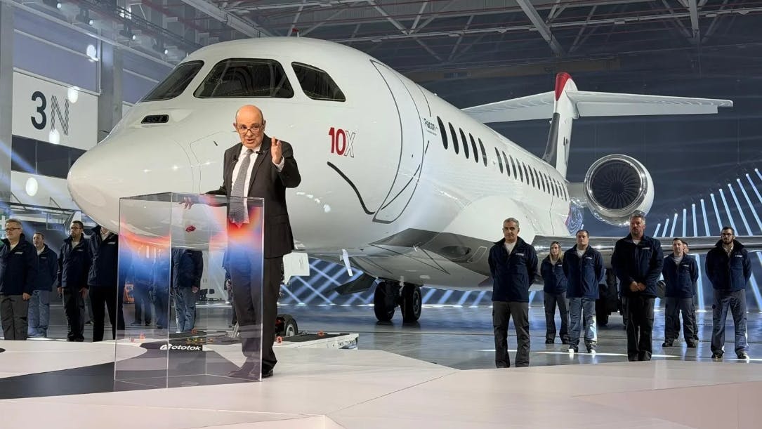 A man wearing a suit standing at a podium and speaking in front of a large white airplane with '10X' printed on the side