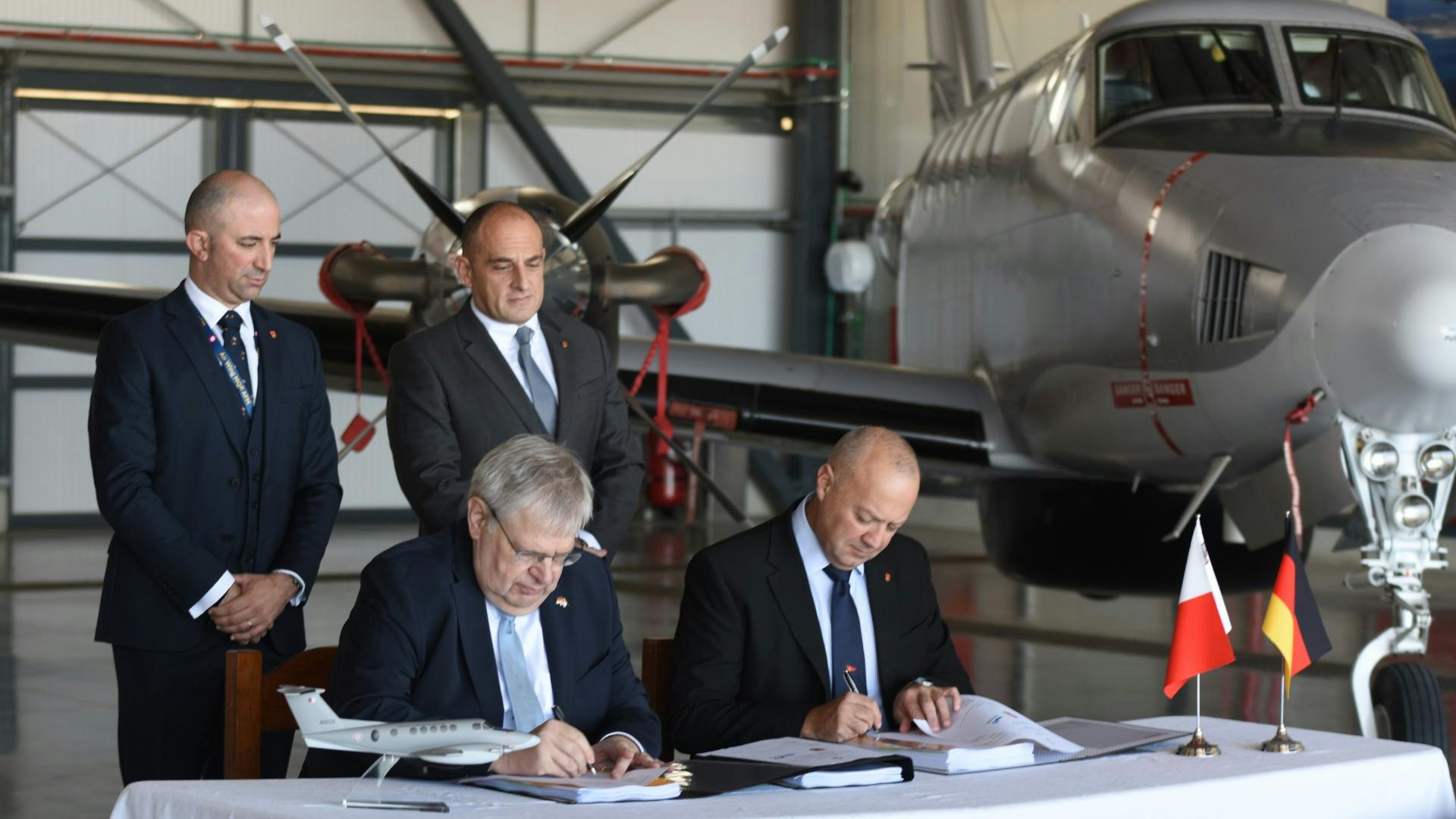 Two men wearing suits standing behind two more men wearing suits sitting down at a table and signing papers, with an airplane parked behind them inside a hangar