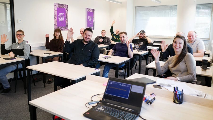 A group of people sitting in a classroom at desks while smiling and waving to the camera