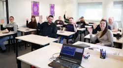 A group of people sitting in a classroom at desks while smiling and waving to the camera A group of people sitting in a classroom at desks while smiling and waving to the camera