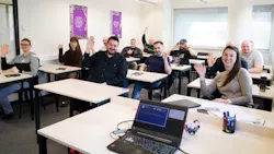 A group of people sitting in a classroom at desks while smiling and waving to the camera A group of people sitting in a classroom at desks while smiling and waving to the camera
