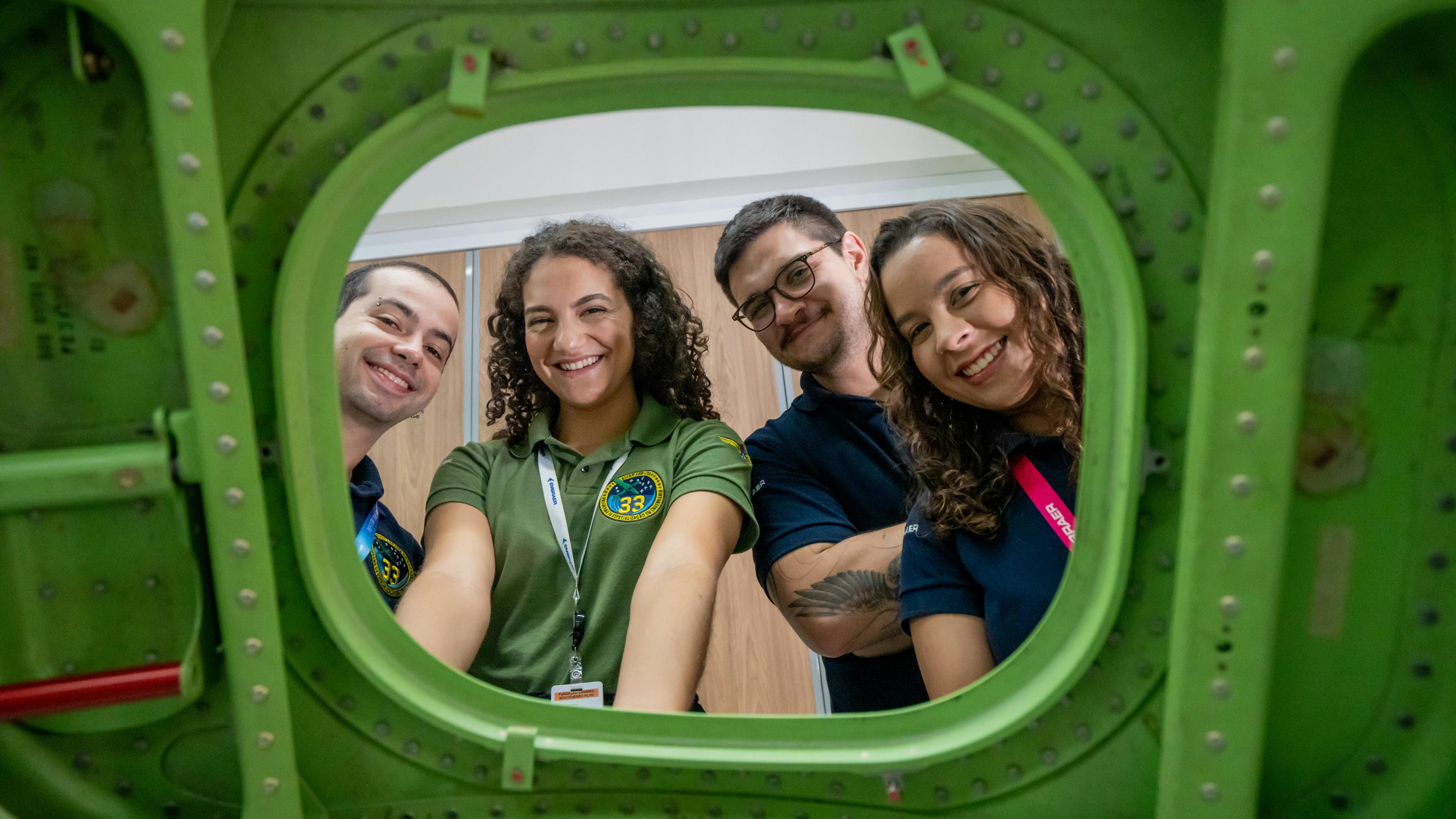 Four people wearing polo shirts and ID badges looking through the window of a green airplane wall smiling at the camera