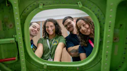 Four people wearing polo shirts and ID badges looking through the window of a green airplane wall smiling at the camera Four people wearing polo shirts and ID badges looking through the window of a green airplane wall smiling at the camera