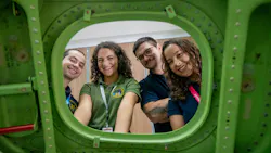 Four people wearing polo shirts and ID badges looking through the window of a green airplane wall smiling at the camera Four people wearing polo shirts and ID badges looking through the window of a green airplane wall smiling at the camera
