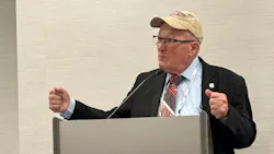 A man wearing business attire and a baseball cap standing at a podium with his arms raised, speaking into a microphone A man wearing business attire and a baseball cap standing at a podium with his arms raised, speaking into a microphone