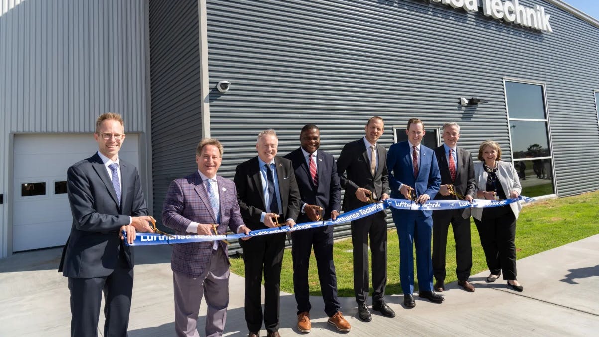 A line of professionals wearing business attire smiling at the camera while standing by a large ribbon during a ribbon-cutting ceremony