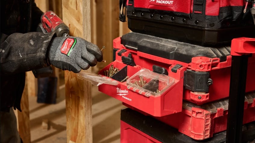 A gloved hand holding a nail next to a toolbox with a compartment on the side filled with nails and screws