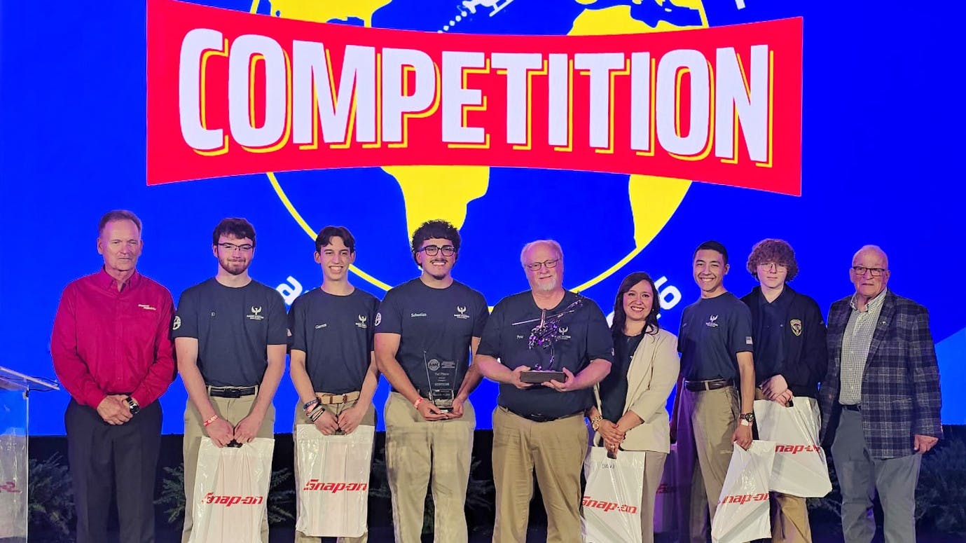 A group of young adults holding gift bags with one holding a trophy, standing in a line between older professionals, under a banner that reads: COMPETITION