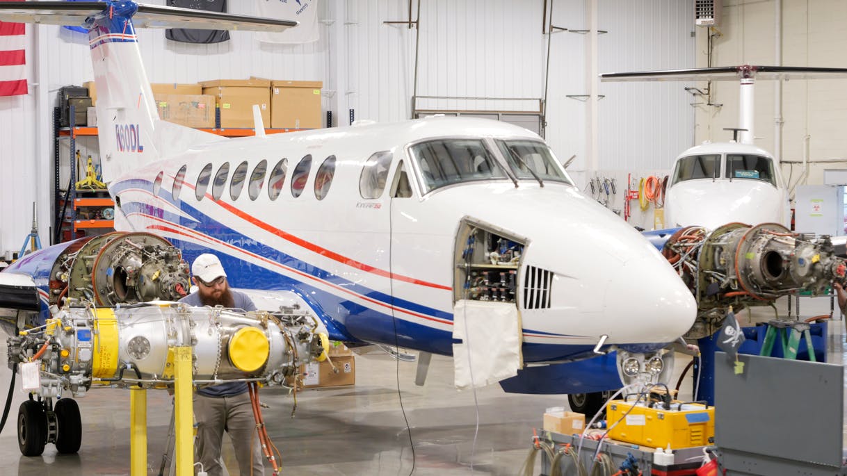 A white airplane with blue and red accents inside a hangar with a side panel open, exposing machinery