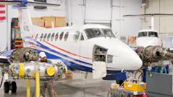 A white airplane with blue and red accents inside a hangar with a side panel open, exposing machinery A white airplane with blue and red accents inside a hangar with a side panel open, exposing machinery