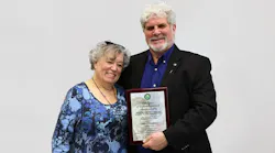 A man and women in professional attire smiling at the camera while standing close to each other, with the man holding a plaque A man and women in professional attire smiling at the camera while standing close to each other, with the man holding a plaque