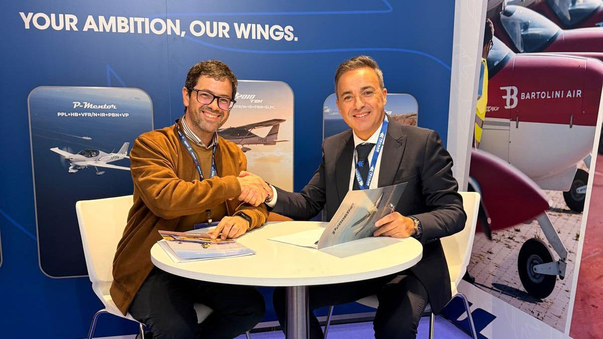 Two men wearing suits sitting at a table and smiling while shaking hands, with a backdrop behind them that reads: YOUR AMBITION, OUR WINGS