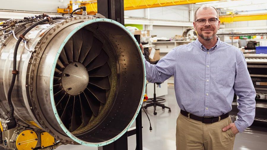 A man wearing khakis and a button-down shirt smiling while leaning on an aircraft engine held in an engine stand