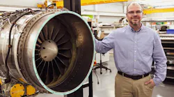 A man wearing khakis and a button-down shirt smiling while leaning on an aircraft engine held in an engine stand A man wearing khakis and a button-down shirt smiling while leaning on an aircraft engine held in an engine stand