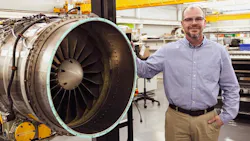 A man wearing khakis and a button-down shirt smiling while leaning on an aircraft engine held in an engine stand A man wearing khakis and a button-down shirt smiling while leaning on an aircraft engine held in an engine stand