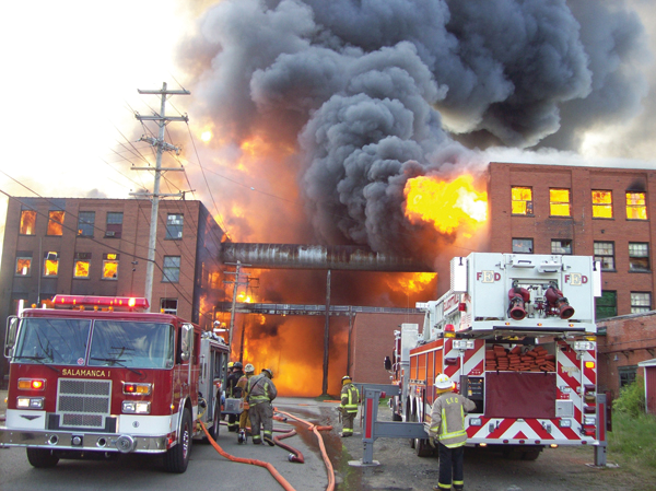 The fire is quickly jumping to an adjoining factory to the right. A mutual aid company is setting up defensive ladder operations, supported by Salamanca&rsquo;s first-arriving engine.