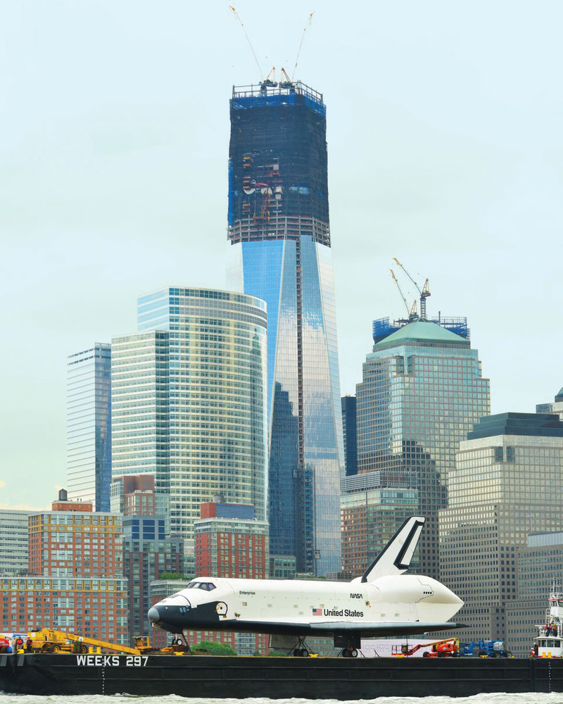 The Space Shuttle Enterprise passes the new World Trade Center Tower 1, where the steel has been completed to its top floor of 104. A tower of some kind, possibly up to 400 feet, may rise from the upper floors next. The building to the right of the 104-story tower with sky cranes visible is the next tower to be rebuilt at the World Trade Center. Building 4 has just reached its scheduled height of 79 stories.