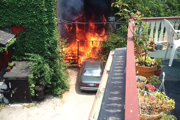 A photo taken from a video shows side C of the fire building prior to the fire department's arrival. The outside deck area is on fire and flames are spreading to a car and the interior of the building through first-floor windows.