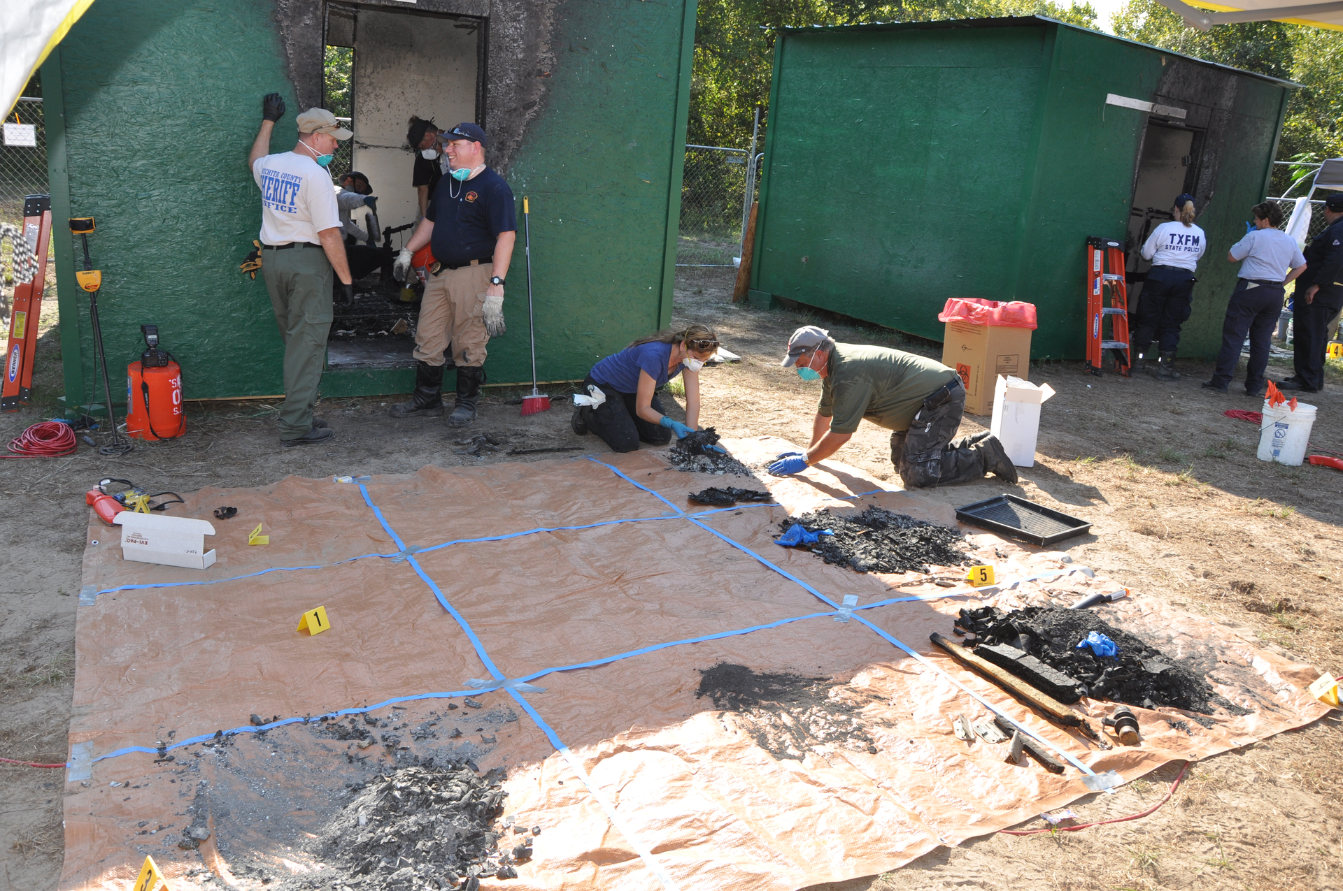 Alan Boyd, left, from the Wichita County, Texas Sheriff&rsquo;s office, discusses the grid method of forensic analysis with Lucas Drago from Temple, Texas Fire Rescue. Kim Burris, Cleburne, Texas Police, and Bill Hollander, with the Bartlesville, Okla. Fire Department, sift through fire debris on a tarp, placing it on a location grid that mirrors the room they are investigating.