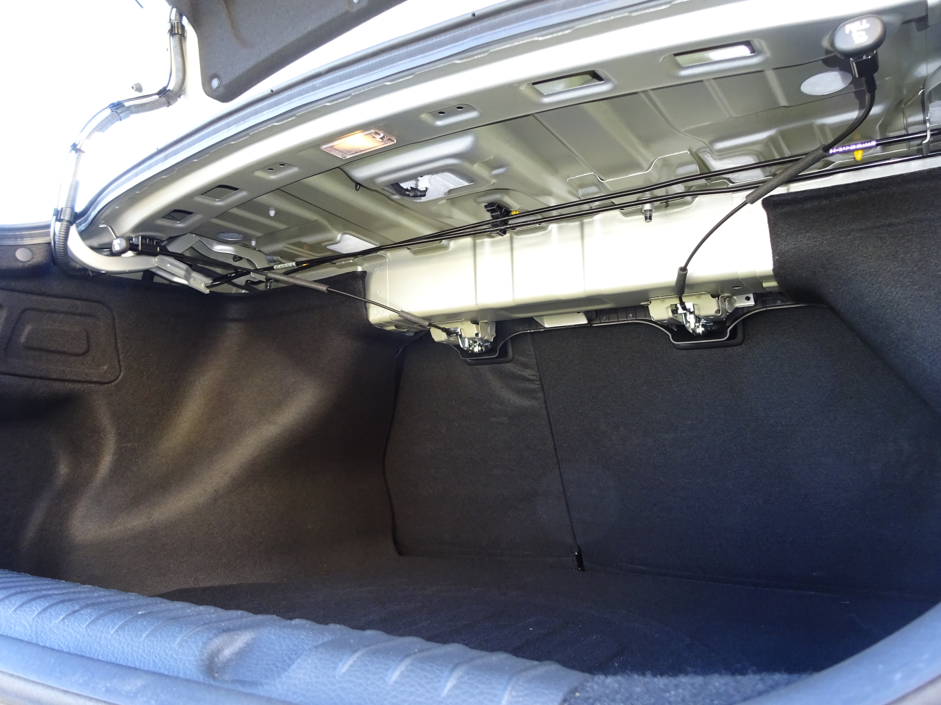 The two black trunk torsion rods of this KIA sedan are visible beneath the rear speaker deck. Note the mild steel mounting bracket for the torsion rods at the left of this image. This is where they can be pried free of the mount and removed.