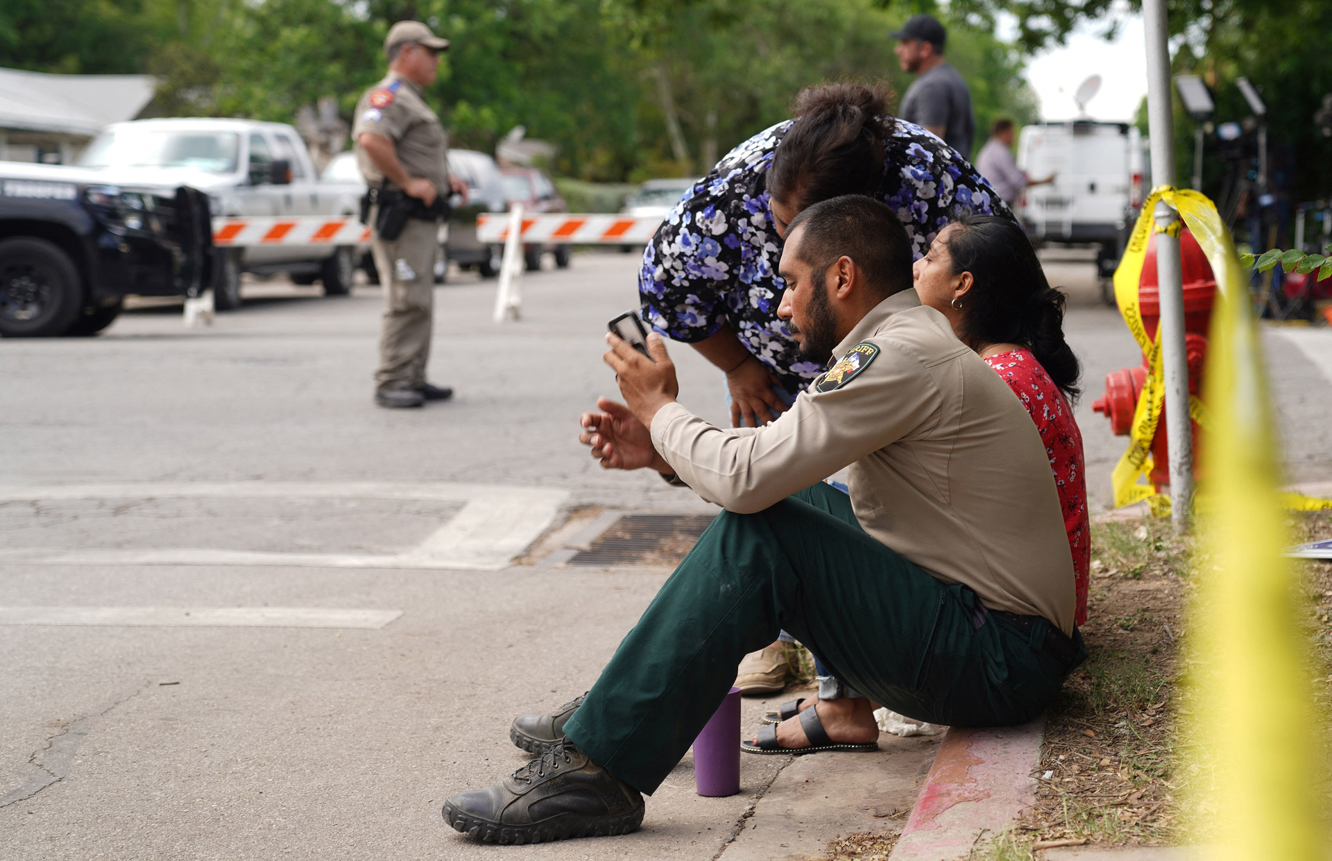 A sheriff checks his phone as he sits on the sidewalk outside Robb Elementary School in Uvalde.