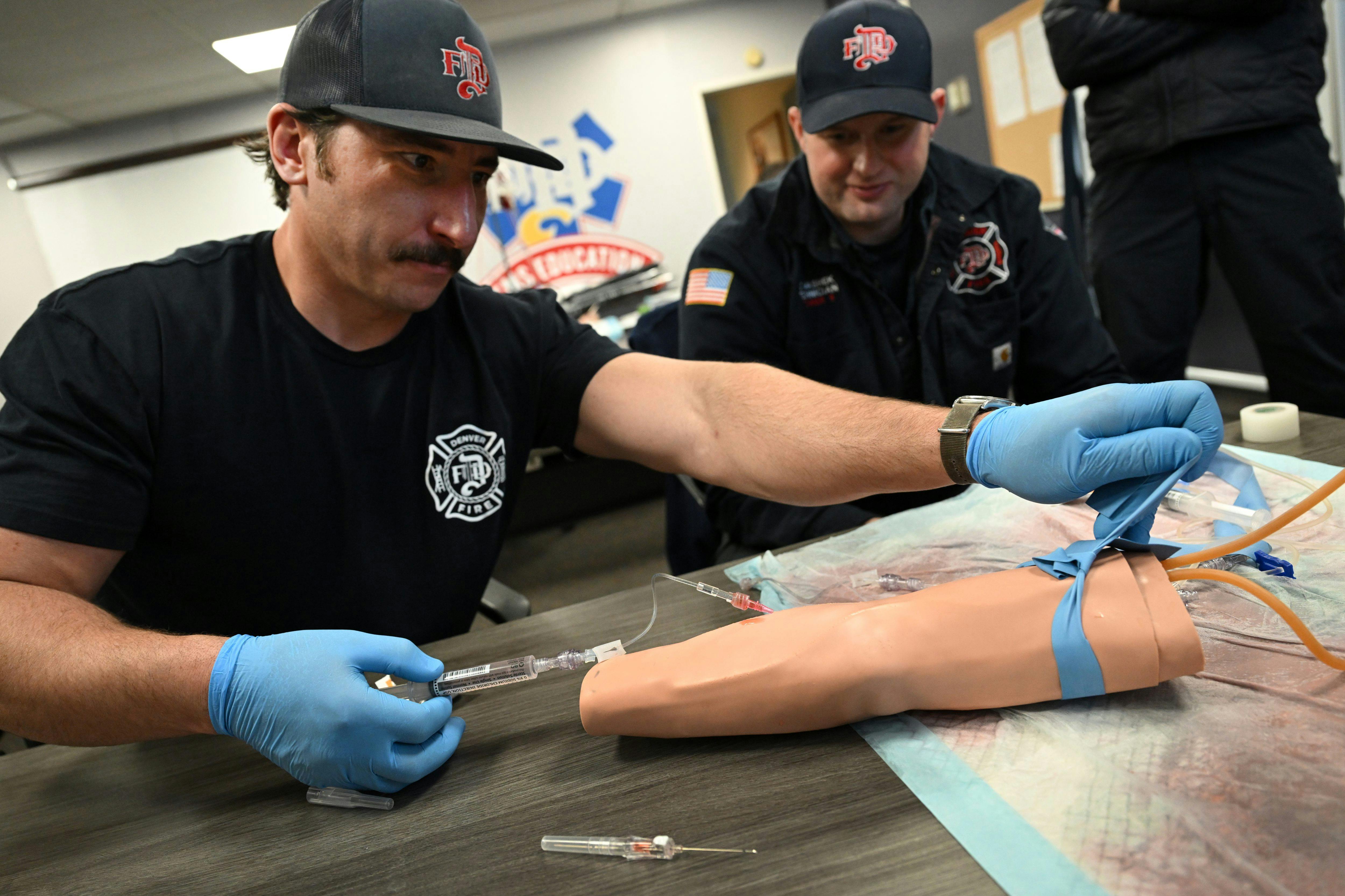 Denver Firefighter Bill Coghill, left, practices placing an IV into a fake arm while hazmat technician Collyn Mashek, right, watches.