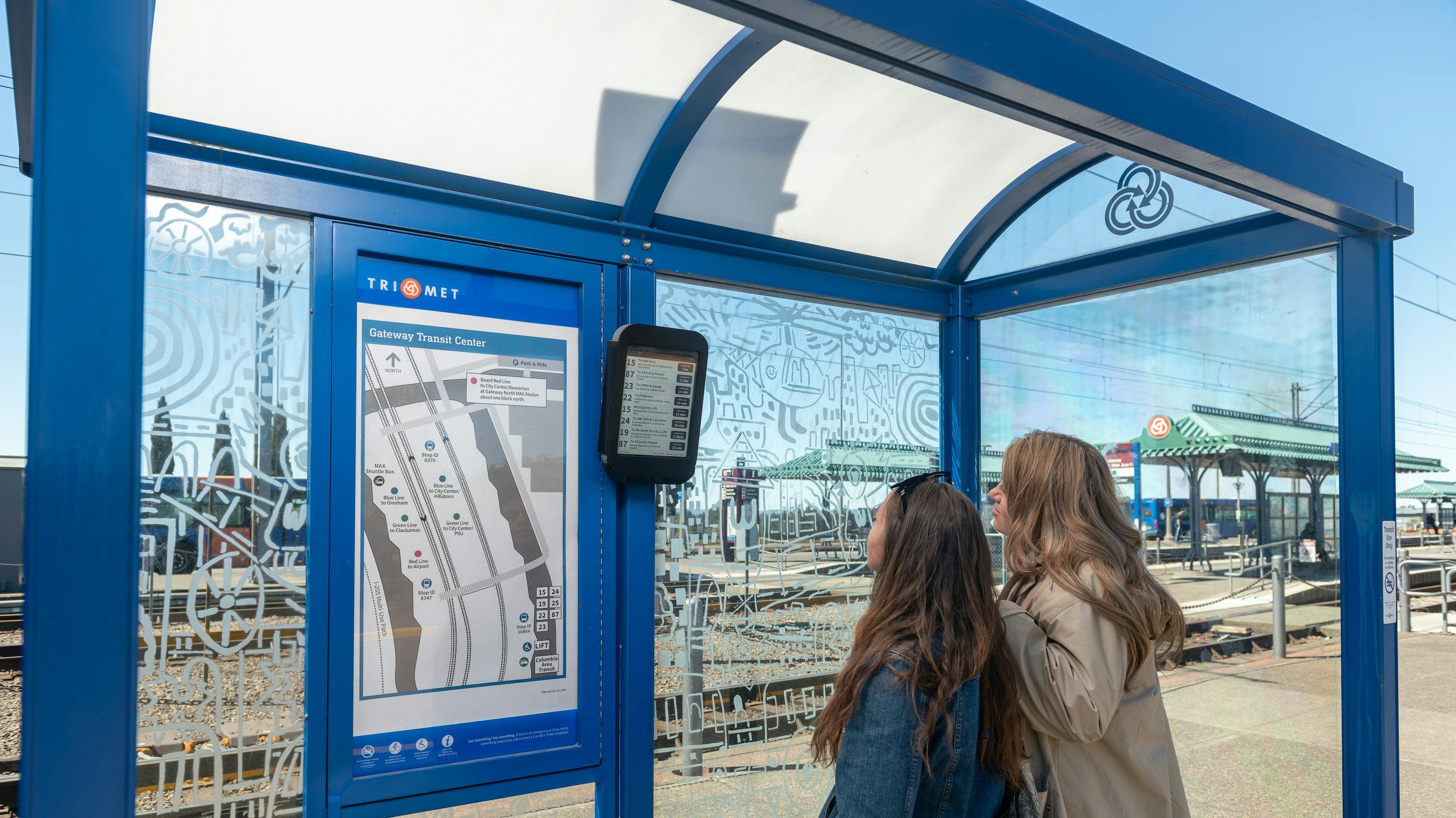 two women stand with back to camera looking at ePaper display in blue bus shelter