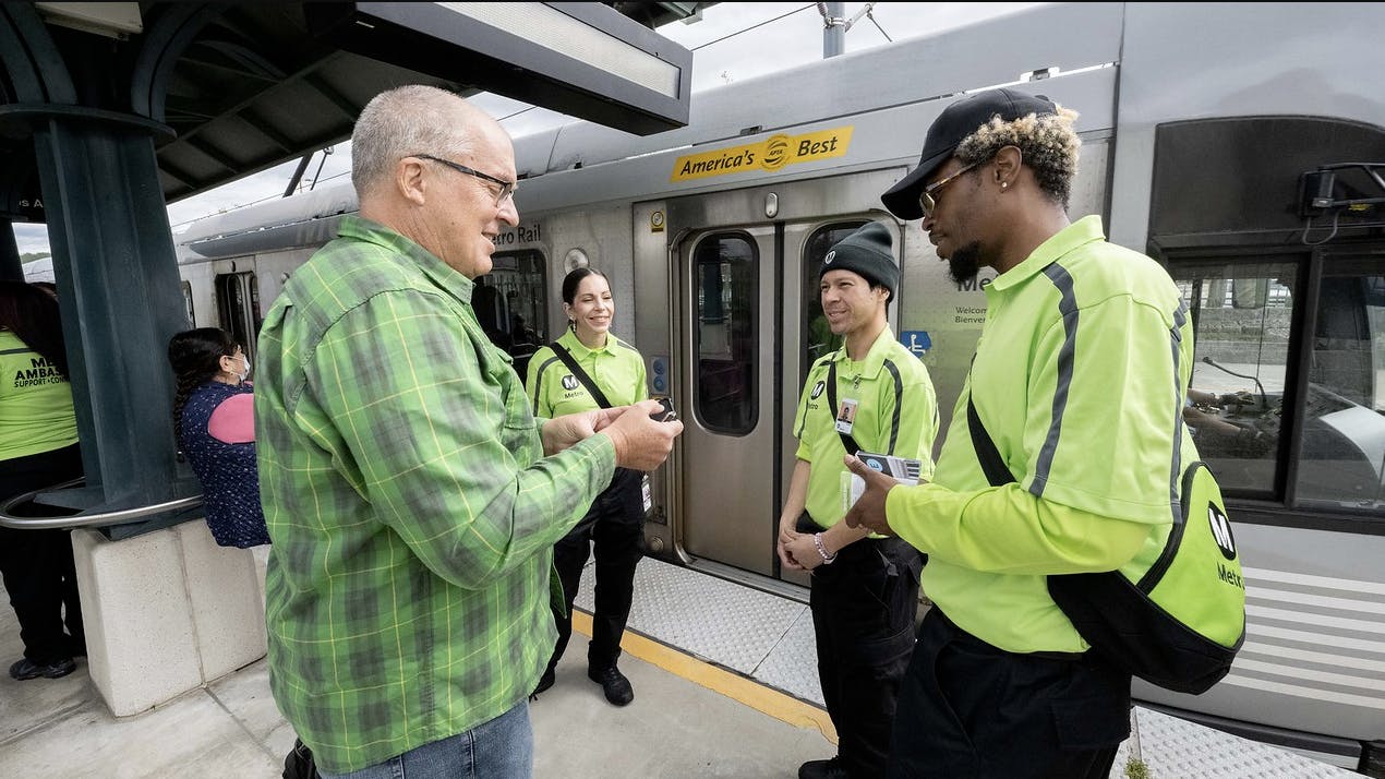 L.A. Metro transit ambassadors dressed in neon green shirts interact with a rider at a rail platform