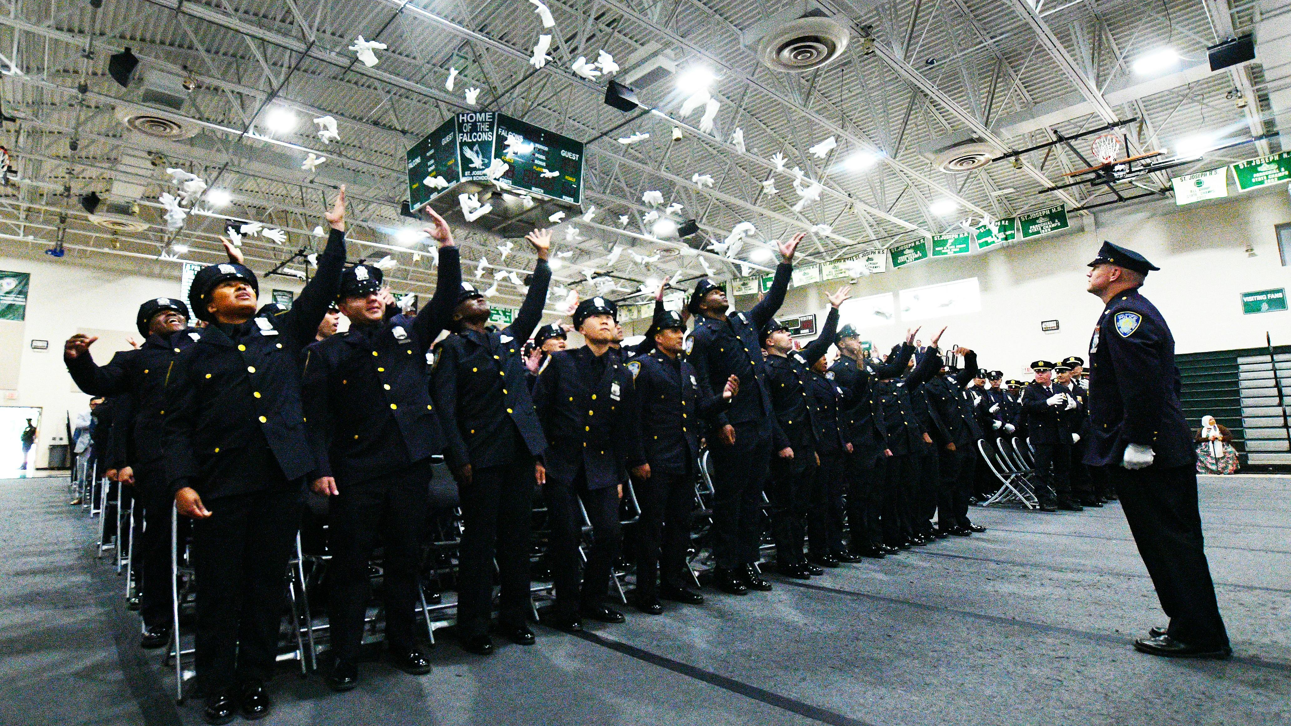 graduating police officers in dress blues stand up and throw their white gloves into the air during their ceremony