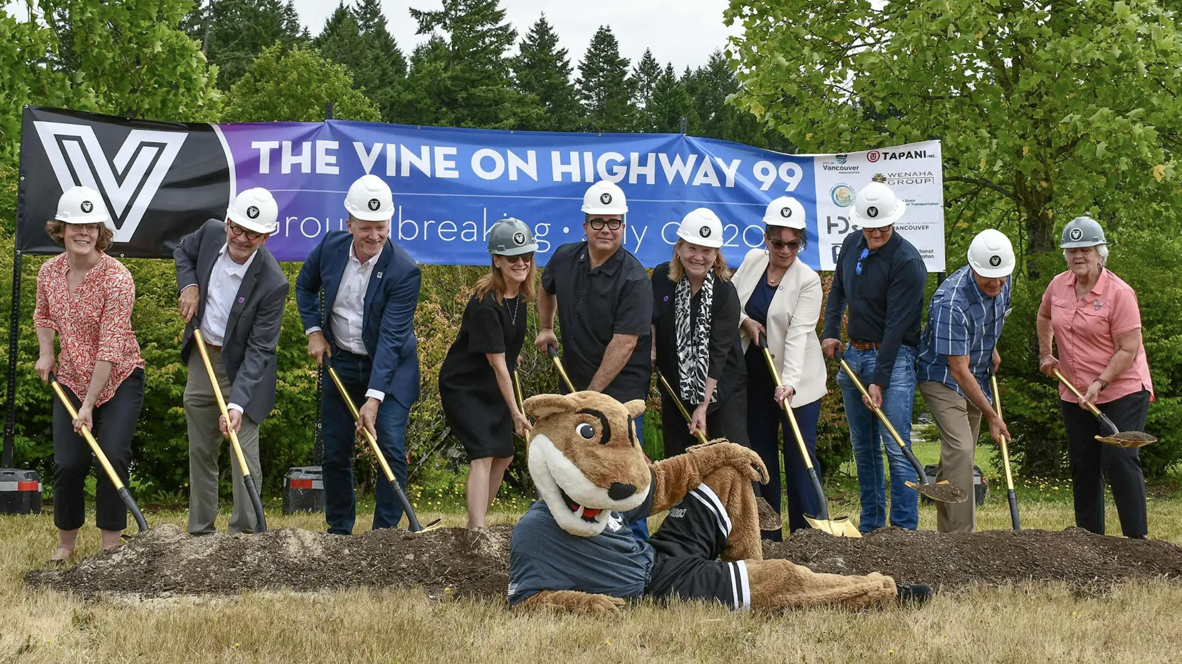 a group of people line up in front of The Vine Highway 99 banner and dig shovels into a pile of dirt. WSU beaver mascot sits in front.