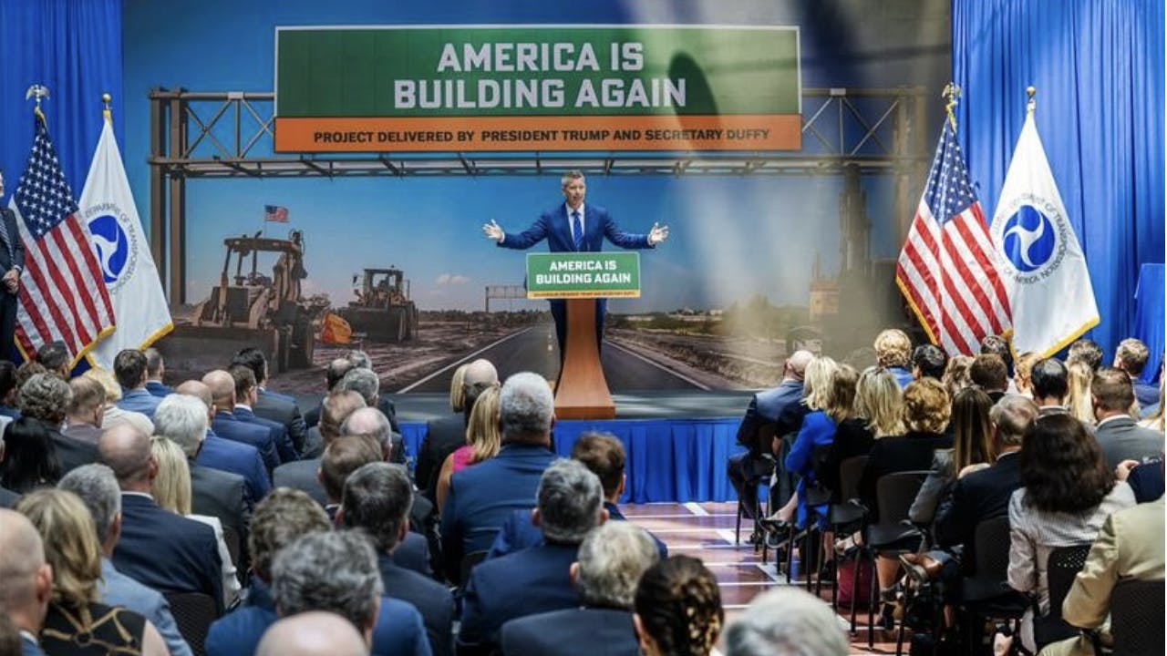 secretary duffy stands at a podium in front of a room full of people with a sign behind him that says america is building again