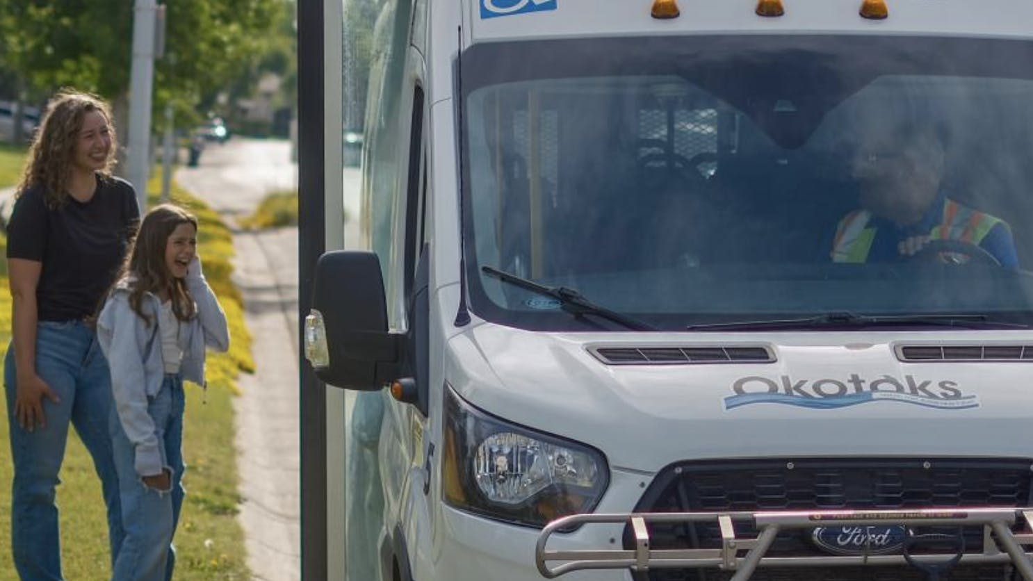 a woman and girl stand on a curb next to an Okotoks Transit cutaway bus